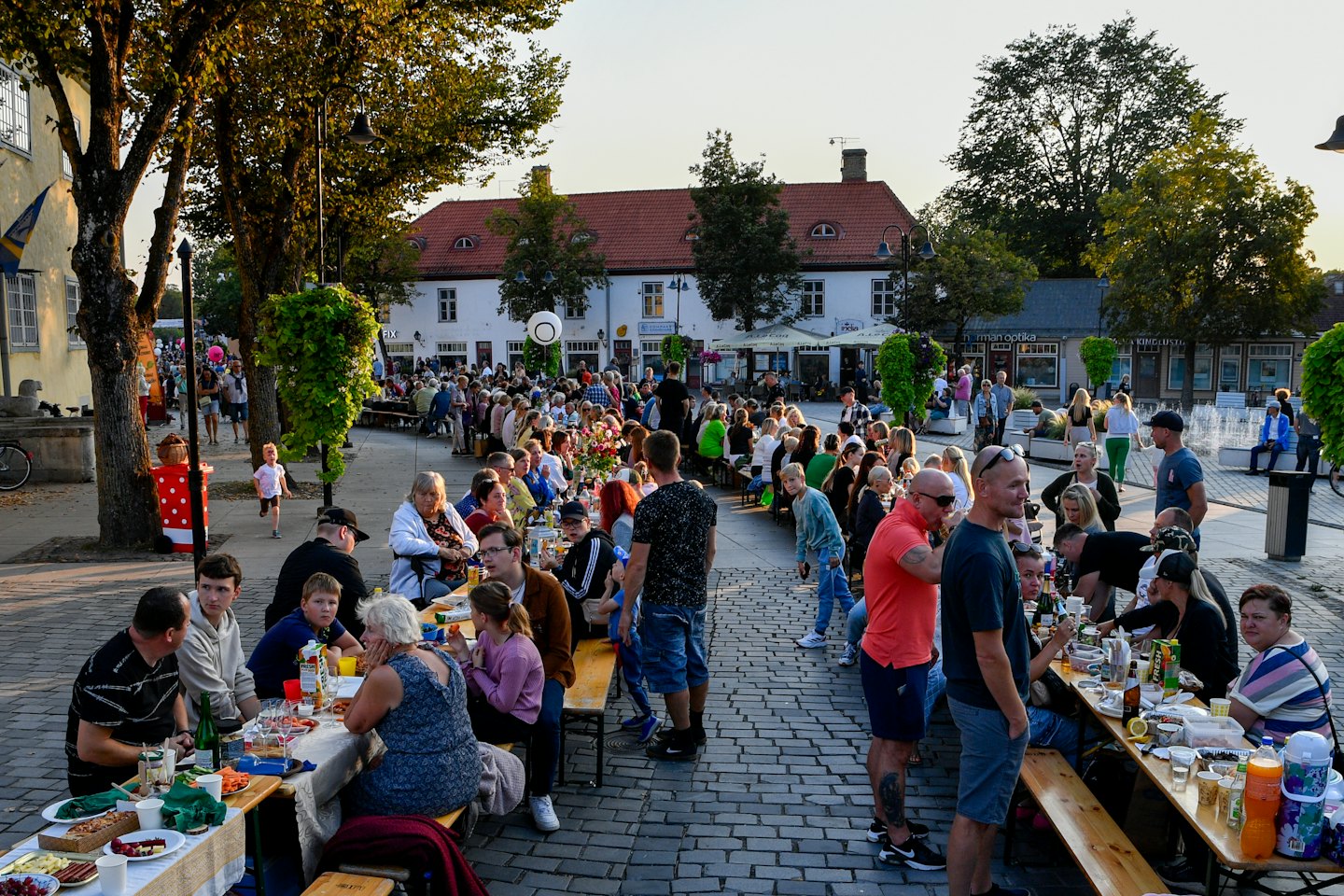 People enjoy food and drinks at tables in a town square at dusk.