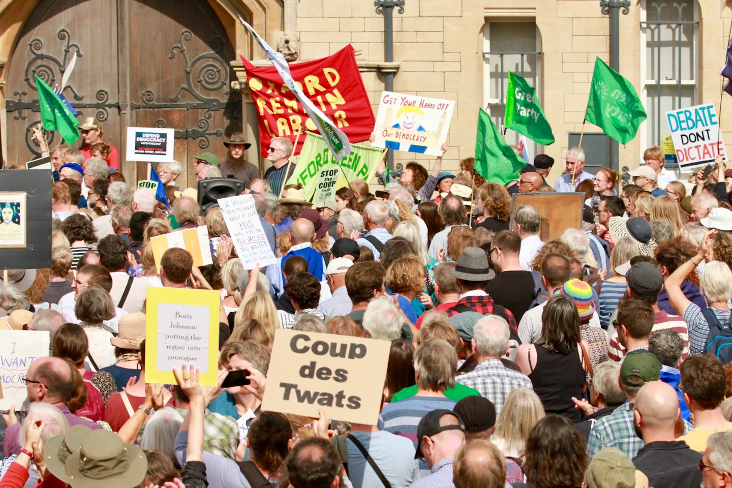 Protesters with signs gather in front of a stone building.